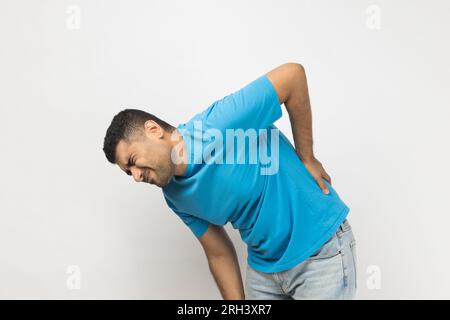 Porträt eines kranken, unrasierten Mannes, der ein blaues T-Shirt trägt, steht und berührt seinen Rücken, hat Probleme mit der Niere, hat ein Trauma, muss behandelt werden. Studioaufnahmen im Innenbereich isoliert auf grauem Hintergrund. Stockfoto
