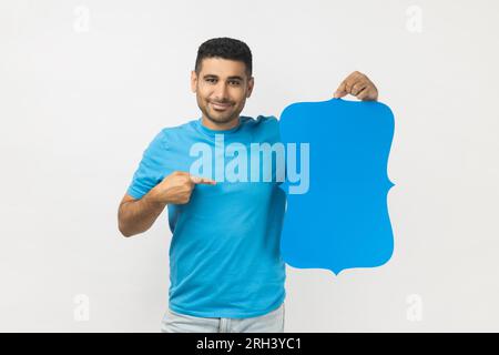 Porträt eines glücklichen, lächelnden, unrasierten Mannes mit blauem T-Shirt, der auf das blaue Sprechblasen-Chat-Symbol zeigt, Platz für Wörter. Studioaufnahmen im Innenbereich isoliert auf grauem Hintergrund. Stockfoto