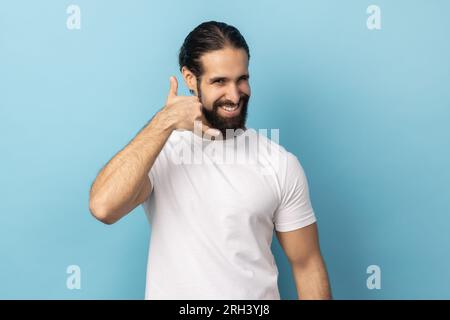 Ruf mich an. Porträt eines Mannes mit Bart, der ein weißes T-Shirt trägt, mit einer Telefongeste spricht und lächelt, flirtet, Kontakt fragt. Innenstudio, isoliert auf blauem Hintergrund. Stockfoto