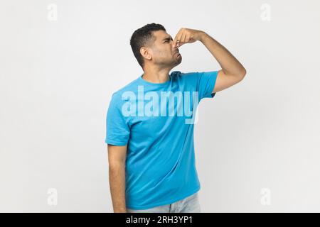 Portrait eines ekelhaften, gutaussehenden, unrasierten Mannes, der ein blaues T-Shirt trägt, steht und die Nase kneift, riecht furchtbar, sein Gesicht dreht. Studioaufnahmen im Innenbereich isoliert auf grauem Hintergrund. Stockfoto
