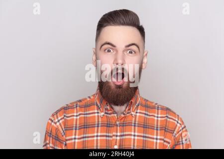 Portrait eines erstaunten, schockierten, bärtigen Mannes, der mit weit offenem Mund in die Kamera schaut, etwas Überraschtes sieht, ein kariertes Hemd trägt. Studioaufnahmen im Innenbereich isoliert auf grauem Hintergrund. Stockfoto