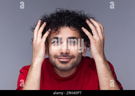 Portrait eines lächelnden, attraktiven, gutaussehenden, bärtigen Mannes mit dunklen Locken, der in den Spiegel schaut, bewundert seine Frisur, trägt einen roten Pullover. Studioaufnahmen im Innenbereich isoliert auf grauem Hintergrund. Stockfoto
