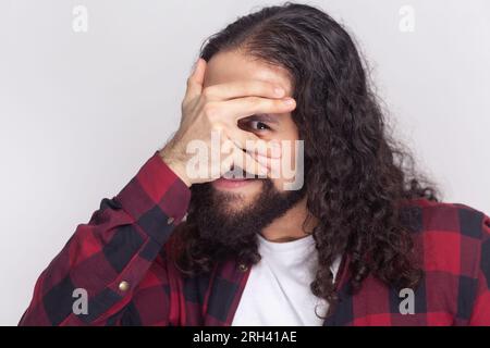 Ein faszinierter bärtiger Mann mit langen Locken im karierten roten Hemd blickt durch die Finger, bedeckt das Gesicht mit Handflächen, versteckt sich, hat Schrecken von etwas. Studioaufnahmen im Innenbereich isoliert auf grauem Hintergrund. Stockfoto