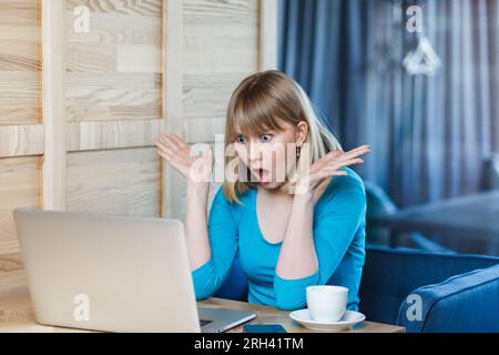 Portrait einer wütenden, schockierten, schönen Frau mit blondem Haar in blauem Hemd, die am Tisch sitzt und an einem Laptop arbeitet, die Hände ausstreckt und fragt, was. Innenaufnahme, Café-Hintergrund. Stockfoto