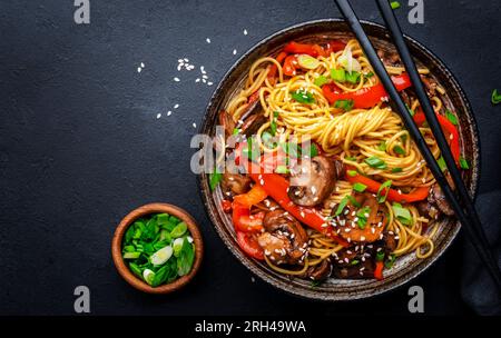 Bratnudeln mit Gemüse umrühren: Rote Paprika, Champignons, grüne Zwiebel und Sesamsamen in Keramikschüssel. Schwarzer Tischhintergrund, Draufsicht Stockfoto