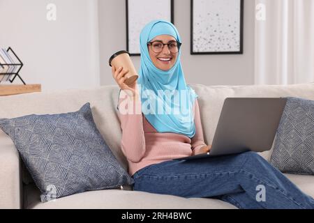 Muslimische Frau mit einer Tasse Kaffee, die ihren Laptop auf dem Sofa im Zimmer benutzt Stockfoto