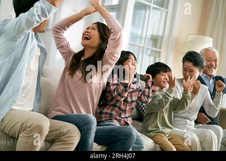 Eine asiatische Familie der 3. Generation, die zu Hause auf dem Sofa sitzt und das Tor und den Sieg feiert, während sie sich die Live-Übertragung des Fußballspiels ansieht Stockfoto
