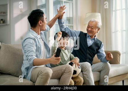 asiatischer Vater Großvater, der zu Hause auf der Couch sitzt und das Tor und den Sieg feiert, während er zusammen im Fernsehen live die Übertragung des Fußballspiels ansieht Stockfoto