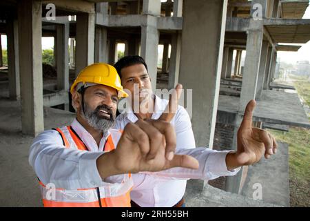 Zwei indische Bauingenieure oder Architekten mit Helm und Weste, die einen Papierentwurf auf der Baustelle halten und über Immobilienprojekte sprechen Stockfoto