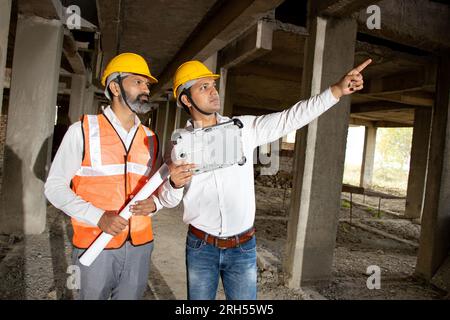 Zwei indische Bauingenieure oder Architekten mit Helm und Weste, die einen Papierentwurf auf der Baustelle halten und über Immobilienprojekte sprechen Stockfoto