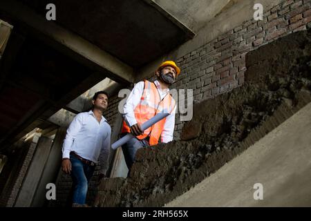 Zwei männliche indische Bauingenieure oder Architekten mit Helm und Weste mit Papierkram, die auf Treppen des Unterbaus laufen und Stockfoto