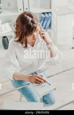 Frau im Büro am Computer. Sie trägt ein weißes Hemd und blaue Jeans, die an einem Tisch sitzen, auf dem ein Laptop und ein Notizblock stehen. Funktioniert in Stockfoto
