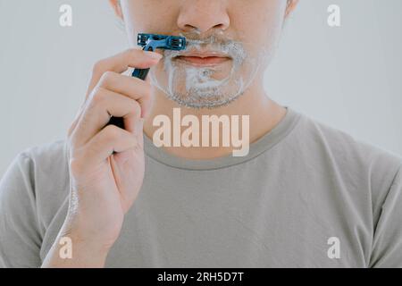 Selektiver Fokus und Nahaufnahme eines Mannes, der ein lässiges, salbeigrünes T-Shirt trägt, rasiert seinen Schnurrbart mit Rasierer und Creme mit flachem Gesichtsausdruck auf IS Stockfoto