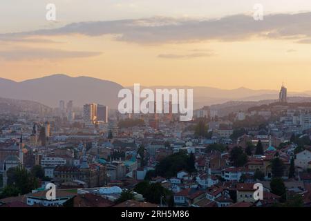 Erhöhter Blick über die Stadt Sarajevo bei Sonnenuntergang mit Apartmentblöcken und Bergen im Hintergrund, Bosnien und Herzegowina, 13. August 2023. Stockfoto