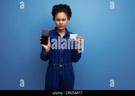 Die junge hispanische Brünette mit flauschigen lockigen Haaren im blauen Jeansanzug zeigt Smartphone und Kreditkartenmockup. E-Business-Konzept Stockfoto