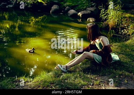 Ein Mädchen in einem Minzkleid am See füttert eine Ente aus der Hand Stockfoto