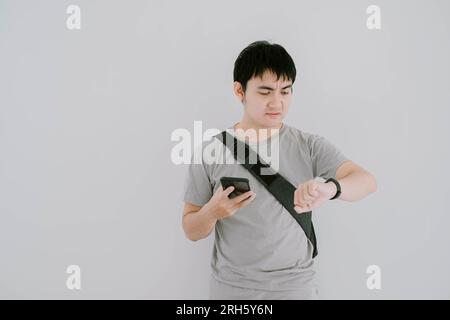 Die selektive Fokusaufnahme eines jungen asiatischen Mannes, der ein lässiges T-Shirt in Salbeigrün, eine blaue Jeanshose und eine Gürteltasche trägt, hält ein Smartphone und schaut auf Smartwat Stockfoto