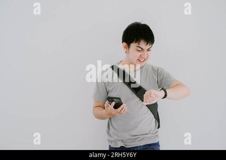 Selektive Fokusaufnahme eines jungen asiatischen Mannes, der ein salbeigrünes lässiges T-Shirt trägt, hält sein Smartphone und schaut auf seine Smartwatch Stockfoto