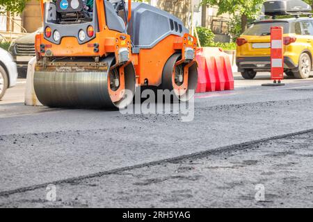 Orangefarbene Straßenvibrationswalze, die an einem Sommertag frischen Asphalt verdichtet. Straßenhintergrund der Stadt. Speicherplatz kopieren. Stockfoto