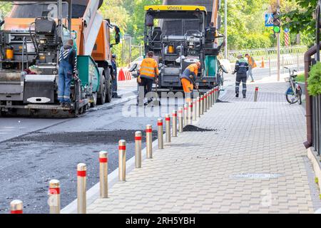 Industrielle Deckenfertiger, die von Straßenbauern betrieben werden, reparieren an einem Frühlingstag eine Straße, die frischen Asphalt auf einer Stadtstraße verlegt. Speicherplatz kopieren. 05. 20. 2023. Kiew, Ukraine. Stockfoto