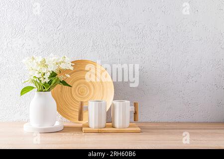 Zwei stilvolle weiße Keramikbecher mit Holzgriff auf einer modernen Arbeitsfläche mit einer Vase und einer Schüssel im Küchenbereich in hellen Farben. Umweltfreundlicher Style Stockfoto