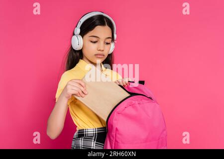 Brünette Schülerin mit kabellosen Kopfhörern, die ein Buch in den Rucksack steckt, pinkfarbener Hintergrund, Student Stockfoto