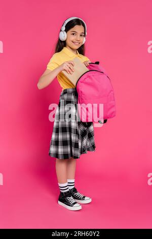 Glückliches Schulmädchen mit kabellosen Kopfhörern, das Buch in den Rucksack steckt, rosa Hintergrund, Student Stockfoto