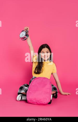 Positives und brünettes Schulmädchen, das mit Rucksack sitzt und den Vintage-Wecker auf Pink hält Stockfoto