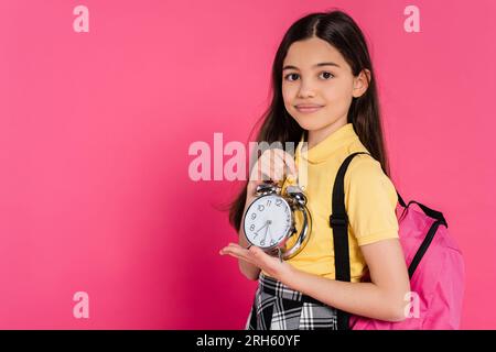 Fröhliches braunes Schulmädchen steht mit Rucksack und hält einen alten Wecker isoliert auf Pink Stockfoto