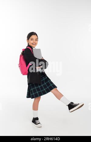 Fröhliches Schulmädchen, das den Laptop hält und in die Kamera schaut, Mädchen in Schuluniform auf weißem Hintergrund Stockfoto