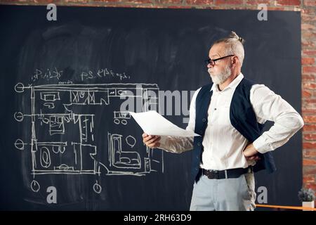 Senior-Mann in stilvoller Kleidung, der architektonisches Design, Zeichnungen, Skizzen mit durchdachtem Gesicht ansieht, Fehler löst Stockfoto