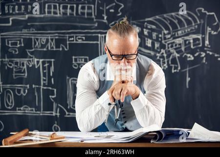 Senior-Mann in stilvoller Kleidung, der architektonisches Design, Zeichnungen, Skizzen mit durchdachtem Gesicht ansieht, Fehler löst Stockfoto