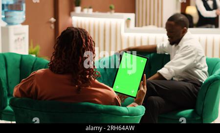 Glückliche Gäste in der Hotellounge halten ein Mock-Up-Tablet mit Chroma Key-grünem Bildschirm und genießen dabei eine Tasse Kaffee. Ein afroamerikanisches Paar wartet auf den Check-in, sitzt auf einem gemütlichen Sofa Stockfoto