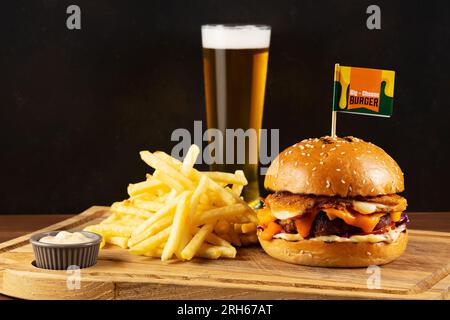 Fast-Food-Konzept. Leckerer Cheeseburger mit gebratenen Kartoffeln und einem Glas Bier auf einem Holztisch. Stockfoto