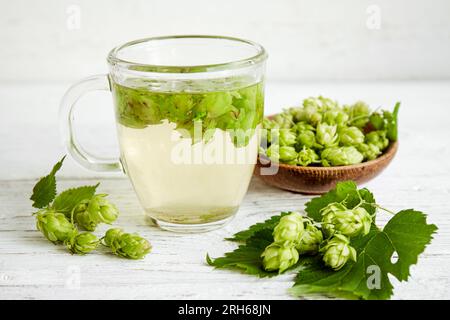 Kräutertee-Getränk aus Humulus lupulus, dem Hopfen oder Hopfen. Hopfenblumen mit Teetasse auf weißem Holz Hintergrund, innen zu Hause. Stockfoto