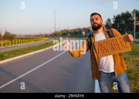 Der Mann trampt am Straßenrand und versucht, das Auto anzuhalten. Er hält Pappe mit Inschrift in der Hand. Stockfoto