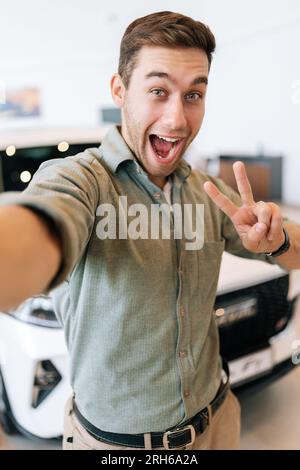 Vertikales POV-Bild eines fröhlichen Käufers, der nach dem Kauf eines neuen Autos ein Selfie mit Siegeszeichen auf dem Smartphone des Autohauses macht. Stockfoto
