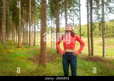 Sportliche Frau trainiert im Freien in einem Wald und hört Musik mit Kopfhörern. Augen zu, sie atmet ein. Stockfoto