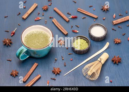 Matcha-Tee in einer blauen Tasse, Milch und Matcha-Teepulver. Gewürze auf dem Tisch. Blauer Hintergrund Stockfoto