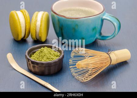 Schneebesen, Bambuslöffel und Matcha-Teepulver in einer schwarzen Schüssel. Einen Makkaronkuchen und eine Tasse Matcha-Tee. Blauer Hintergrund Stockfoto