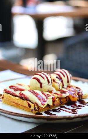 Belgische Waffeln mit Eis, Beerensauce und frischen Obstscheiben auf einem Teller auf einem Restauranttisch. Vertikales Bild von Vollkornmehl belgischer Waffel Stockfoto