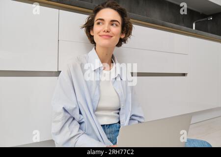 Junge Frau, die auf dem Boden lernt. Mädchen mit Laptop arbeitet von zu Hause aus, schreibt mit dem Computer, lächelt und fühlt sich kreativ. Stockfoto