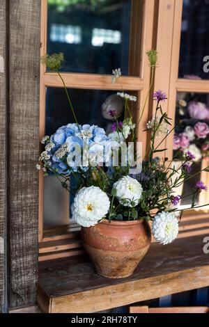 Ein rustikaler Strauß aus blauen Hortensien, weißen Chrysanthemen und Dahlien sowie Zichorien in einer braunen Flasche auf einem Holztisch als Dekoration der Veranda Stockfoto