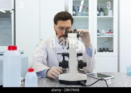 Konzentrierter und seriöser männlicher Arzt, Wissenschaftler arbeitet in einem Labor, sitzt an einem Tisch und schaut sich ein Mikroskop an, studiert, recherchiert, macht eine Analyse. Stockfoto