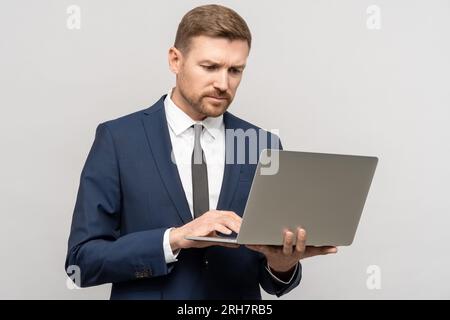 Ein seriöser Geschäftsmann im Anzug, der auf einen Notebook-Bildschirm auf grauem Hintergrund schaut. Stockfoto