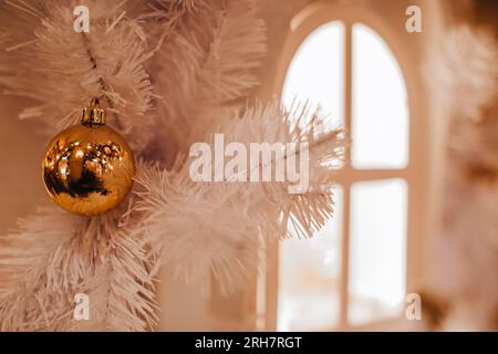 Goldene glänzende Weihnachtskugel, die auf dem weißen Weihnachtsbaum hängt. Magische Winterdetails mit goldenen Bokeh-Lichtern auf dem Hintergrund. Neujahrsferien Wallpa Stockfoto