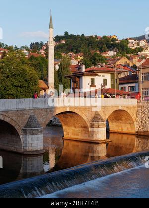 Brücke über den Fluss Miljacka mit Restaurant Inat Kuca und Minarett dahinter in Sarajevo, Bosnien und Herzegowina, 14. August 2023. Stockfoto