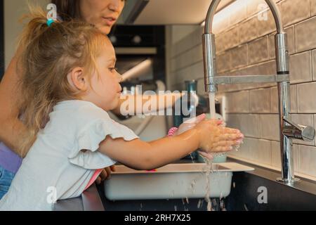 Das kleine Mädchen wäscht sich die Hände in der Spüle, während die mutter hilft, damit sie nicht umfällt. Hygiene und Sauberkeit sind moderne Zivilisation Stockfoto