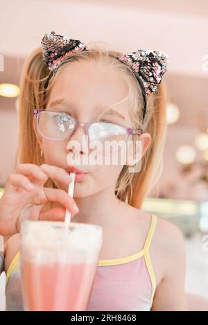 Ein hübsches Mädchen, sieben Jahre alt, trinkt Erdbeermilchshake durch einen Strohhalm in einem Café. Portrait des glücklichen Mädchens, das einen Cocktail trinkt Stockfoto