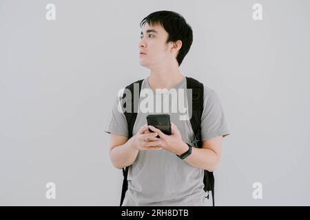 Ein junger asiatischer Mann, der ein lässiges T-Shirt in Salbeigrün, eine Smartwatch und einen Rucksack trägt, sucht auf der linken Seite nach Wegbeschreibungen, während er das Smartphone hält Stockfoto